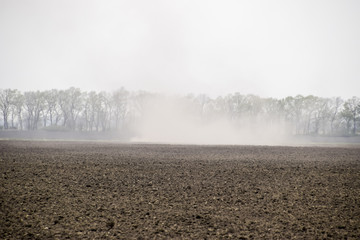 The tractor harrows the soil on the field and creates a cloud of dust behind it