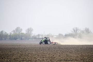 The tractor harrows the soil on the field and creates a cloud of dust behind it