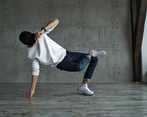 Teenage boy in white sweater and baseball cap, jeans and sneakers makes breakdance in dance studio....