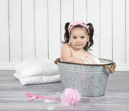 Sweet, toddler girl in  pink headband sits in bubble bath in tiny, metal wash tub 