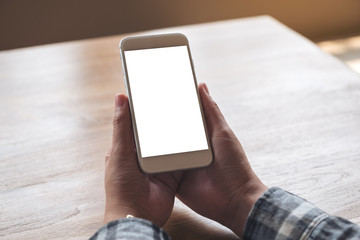 Mockup image of hands holding white mobile phone with blank screen on wooden table in cafe