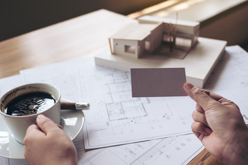 A hand holding and showing an empty business card with an architecture model and shop drawing paper on table in office