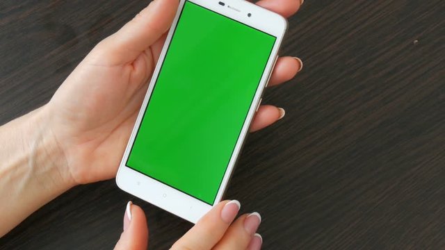 Female hands with beautiful French manicure take a white smartphone with Green Screen. Using Smartphone,Holding Smartphone with Green Screen on a stylish black wooden table