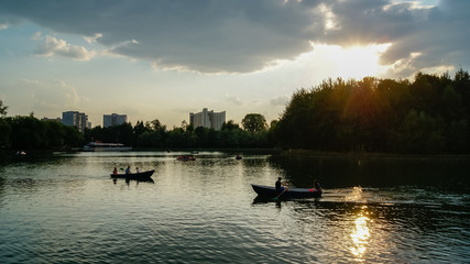 People ride a boat and catamarans on the lake in city park, green trees and ferris wheel on the background