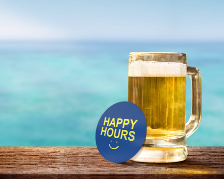 Happy Hour Concept for Bar, Cafe or Hotel Resort to Promote a Special Offer to Customer. Glass of Beer on Wooden Table with Smiley Face and Text on Note. Relaxation on the Seaside