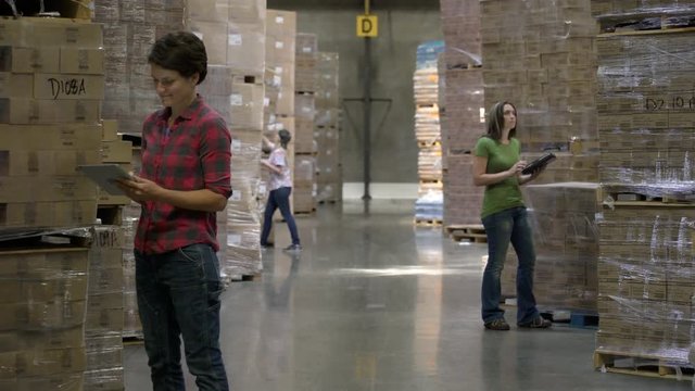 Three female colleagues standing by stock on pallets, writing on clipboards.