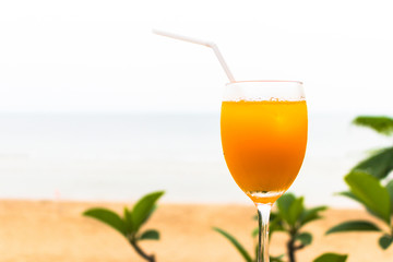 Glass of orange juice with backdrop green leaves and sand beach and sea background.