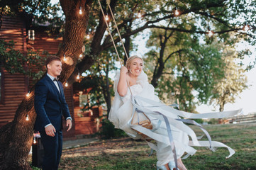 A stylish bridegroom rolls the bride on a swing, against the backdrop of beautiful nature and green grass. Merry time at the wedding. Funny newlyweds are fooling around. Film effect.