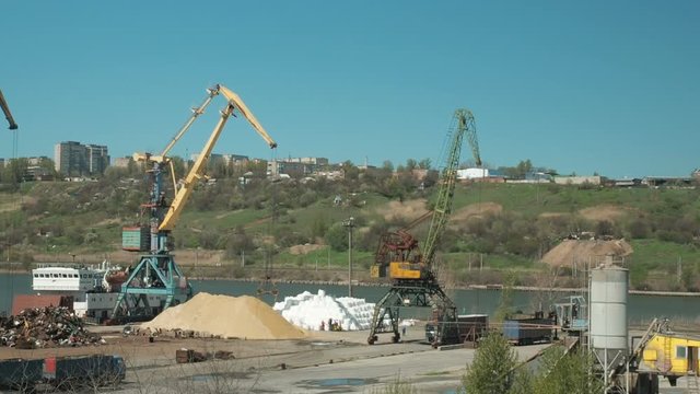 Landscape of tugboats and cranes in shipyard in cargo port. The crane moves sand brought by trucks in a cargo terminal. A large bucket is picking up sand.