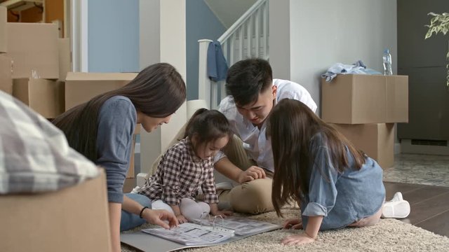 PAN of Asian family with little children sitting on floor on empty living room of new house and looking through folder with furniture layout ideas