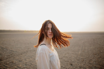 Fototapeta na wymiar Beautiful brunette lady in soil field at sunset. Happy beautiful woman in meadow. Beautiful carefree woman in fields. Portrait of beautiful young woman. Hair blowing in the wind