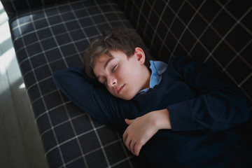 A boy teenager with closed eyes in a blue shirt lies on a blue couch in a white cage and sleeps. The sun's rays fall on the boy's face.