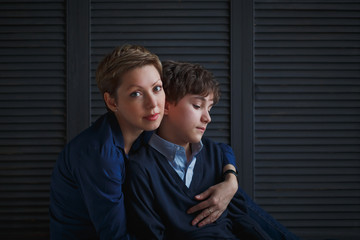 Happy young mom with short hair hugs her teenage son and smiles. The boy looks down with sad eyes.