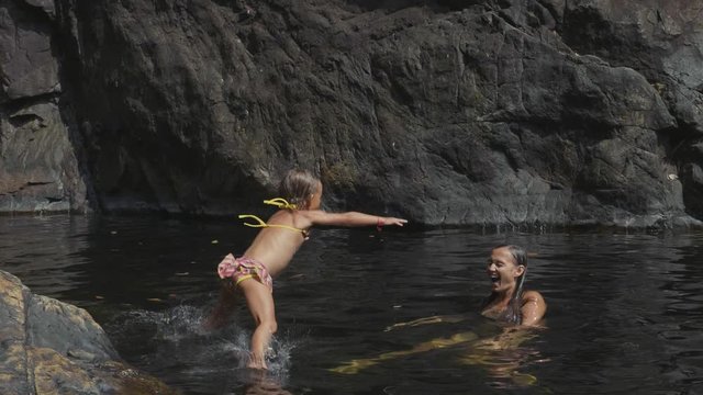Little cute girl jumping in water of mountain lake to her mother