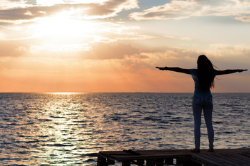 Fototapeta premium A young slender girl stands on a wooden bridge (fishing bridge) spreading her hands over the sea and looking at the sunset (dawn) of the sun. Life and freedom. 