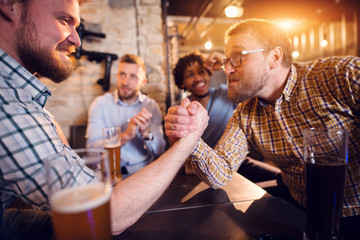 Prepared and focused male friends looking each other while having arm wrestling challenge at the local bar and other supporting.