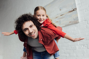 Obraz premium happy father piggybacking cute little daughter and smiling at camera at home