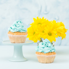 Cupcake with blue cream decoration on stand and bouquet of yellow chrysanthemum in glass vase on blue pastel background.