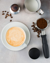 The cup of latte or cappuccino coffee with milk. Background with tools for making fresh boiled coffee: a glass of milk, coffee beans, a horn from a coffee machine.