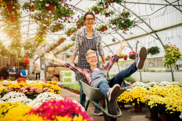 Two adorable joyful florist women having fun with cart for a break in the greenhouse.
