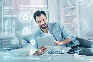 Relaxing alone. Cheerful young emotional actor smiling while looking at the screen of a modern tablet and relaxing on the bed at home