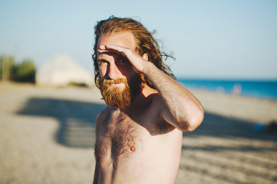 Man with long hair and a beard standing on beach shielding his eyes from the sun