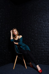 Handsome brunette girl wear on black and green jacket in red high heels, sitting and posing on chair at studio against dark brick wall. Studio model portrait.