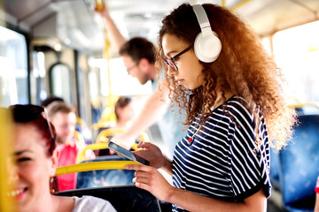 Young beautiful woman is listening to music on her phone while wearing a headset and taking a bus...