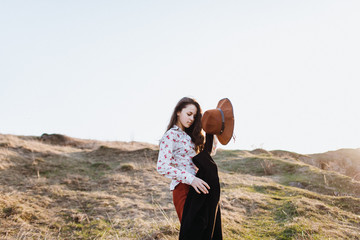 Naklejka premium Beautiful girl in brown pants and with a hat walks to mountain places with a young spring grass. traveler hipster is holding hat in sunset. travel concept