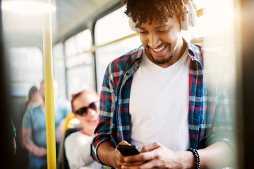 A young joyful man is smiling as he uses his phone and listens to music while traveling by bus.