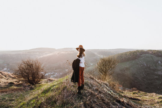 Beautiful girl in brown pants and with a hat walks to mountain places with a young spring grass. traveler hipster is holding hat in sunset. travel concept. View from behind