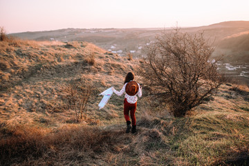 Beautiful girl with a map, binoculars and hat on top of the mountains walking at sunset. Soft focus. Back view