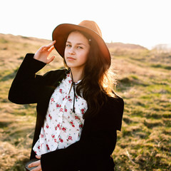 Beautiful girl in a black coat, brown pants and with a hat walks to mountain places with a young spring grass. traveler hipster holding hat in sunset. Wearing stylish fall outfit. close-up portrait