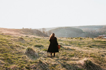 Beautiful girl in a black coat, brown pants and with a hat walks to mountain places with a young spring grass. traveler hipster holding hat in sunset. Wearing stylish fall outfit. rear view