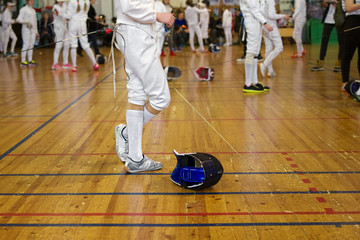 Girl participant in the fencing competition on swords is in the center of gym holding a sword in her hand, waiting for next battle. Fencing mask is on the floor