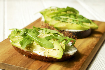 Vegan sandwich, rye bread toast, avocado, vegenaise eggless mayonnaise sauce, sesame seeds seasoning, arugula rucola leaves white wood texture table. National avocado day concept. Close up, background
