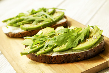 Vegan sandwich, rye bread toast, avocado, vegenaise eggless mayonnaise sauce, sesame seeds seasoning, arugula rucola leaves white wood texture table. National avocado day concept. Close up, background