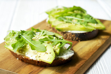 Vegan sandwich, rye bread toast, avocado, vegenaise eggless mayonnaise sauce, sesame seeds seasoning, arugula rucola leaves white wood texture table. National avocado day concept. Close up, background