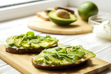 Vegan sandwich, rye bread toast, avocado slices, vegenaise sauce & raw arugula. Toasted sourdough, eggless mayonnaise, wooden table, whole & halved. National avocado day concept. Close up, background.