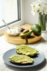 Vegan sandwich, rye bread toast, avocado, whole & halved, vegenaise sauce. Toasted sourdough, eggless mayonnaise, wood slice, daffodil flower vase. National avocado day concept. Close up, background.