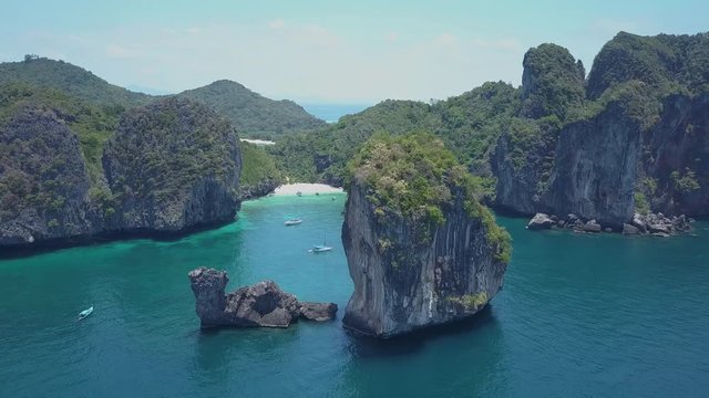 AERIAL: Circling around behemoth limestone cliff overlooking beautiful sandy bay on paradise island. Spectacular shot of limestone karst landscape surrounding tranquil sandy beach and anchored boats.