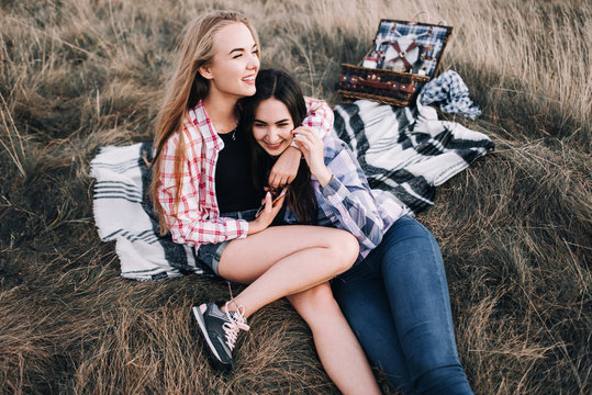 Two beautiful girls in colored shirts sitting on the grass on the hillside. Watch the sunset, drinking wine, hug, happy. picnic