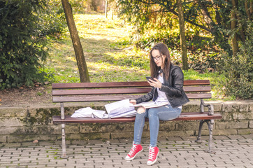 Young beautiful school or college girl with glasses sitting on the bench in the park reading the books and study for exam, calling her friend on the cell phone to ask for help about syllabus
