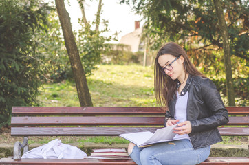 Obraz premium Young beautiful school or college girl with eyeglasses sitting on the bench in the park reading the books and study for exam