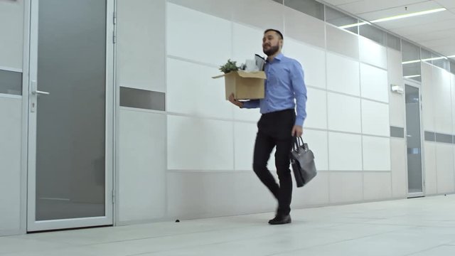 Asian businessman walking with briefcase through hallway and carrying cardboard box filled with personal belongings while moving in new office