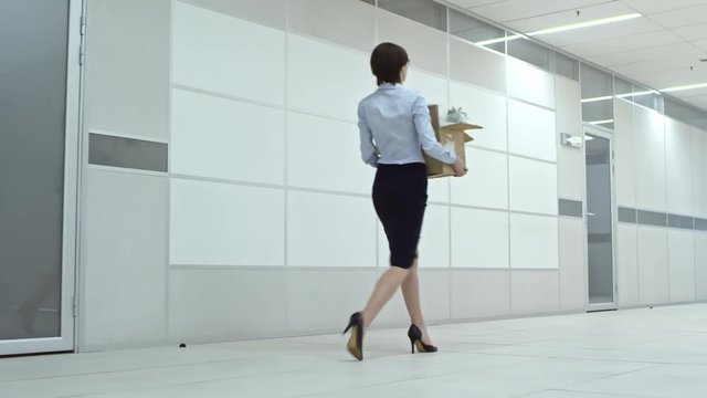 Businesswoman walking through hallway and carrying cardboard box filled with personal belongings while moving in new office