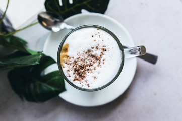 A cup of Hot cappuccino latte coffee with cinnamon sprinkles on top on wooden white dinner table with a green leaf in a  background