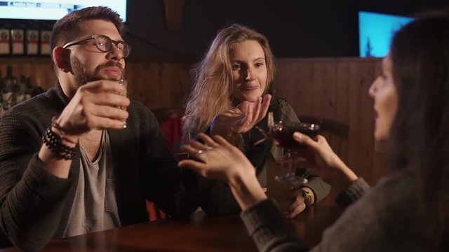 Relaxed man and women are having fun in cafe in evening. They are holding glasses with drinks and chatting joyfully in dark hall