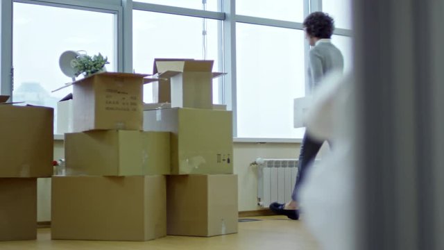 Tracking shot of young businesswoman walking through new office with stacks of cardboard boxes, talking on cell phone and sitting on window sill