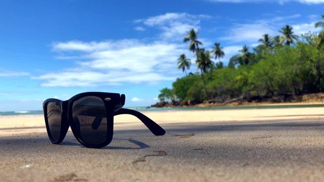 Pair of sunglasses resting under the shadows of palm trees blowing in the wind on a remote tropical island beach